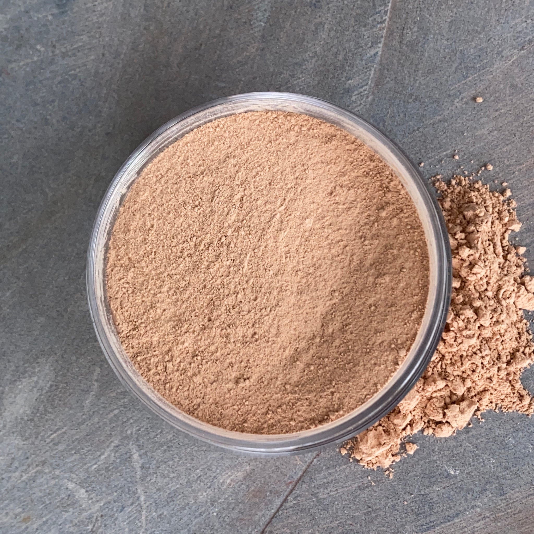 A jar of mineral powder foundation with a small amount of powder gently spilled beside it, symbolizing the purity and mindfulness behind Coastal Classic Creations' makeup products.