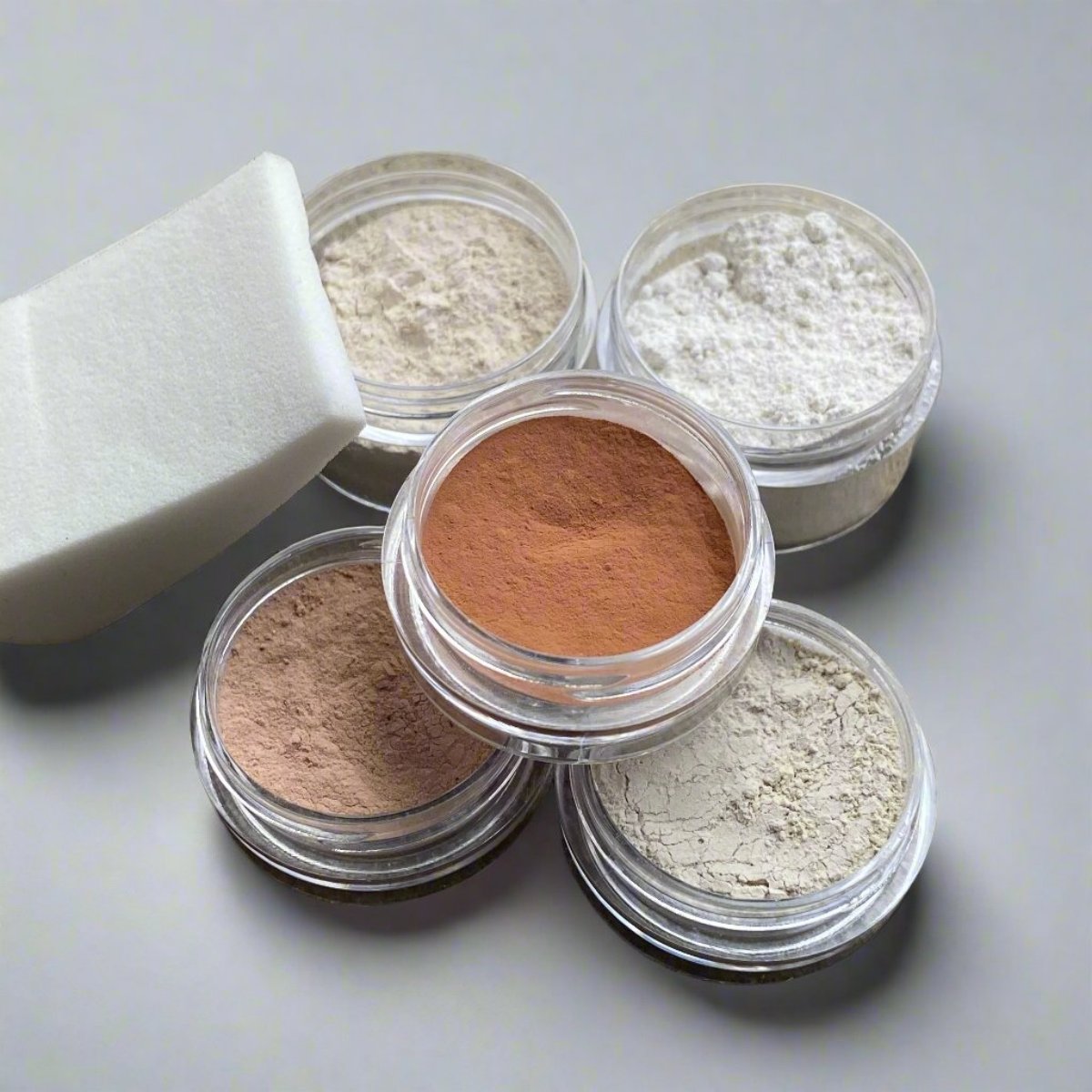 A collection of cosmetic containers including a wedge applicator, foundation, concealer, base primer, and translucent natural mineral powders.